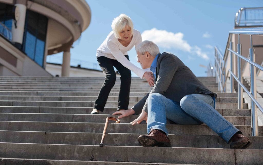 An elderly woman assisting an elderly man who has fallen on the stairs due to balance issues